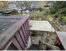 Blick von einer Holztreppe auf einen Garten mit Laub und mehreren kleinen Bauwerken, darunter ein Wellblechdach und ein Gewächshaus. Im Hintergrund sind weitere kleinere Gebäude sowie Bäume und Wohnhäuser zu sehen.