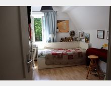 Kleines Schlafzimmer mit Holzboden und einem Einzelbett, das mit einer Patchwork-Decke bedeckt ist. An der Wand hängen Bilder und eine runde Uhr, das Fenster mit blauen Vorhängen bietet Ausblick auf Bäume und Tageslicht. Auf dem Bett stehen mehrere Stofftiere, daneben ein Schreibtisch mit Hocker.