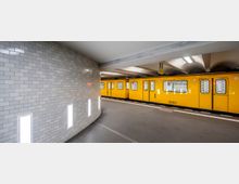 U-Bahn-Station mit hellgrauen, gefliesten Wänden und vertikal eingebauten Leuchten; im Hintergrund steht ein gelber U-Bahn-Zug auf einem Gleis unter einer gewölbten Decke mit modernen Leuchtstofflampen.