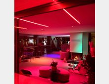 Ein moderner Innenraum mit gemütlicher Lounge-Atmosphäre, beleuchtet durch rotes und violettes Stimmungslicht. Der Raum verfügt über eine Sitzecke mit einem Kamin, umgeben von Möbeln und Dekorationen, sowie Hochtische im Hintergrund.