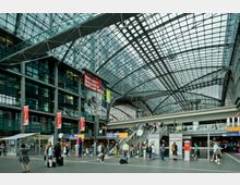 Großer, moderner Bahnhof mit einer Glasdachkonstruktion, mehreren Ebenen und Rolltreppen. Im Vordergrund sind Fahrgäste mit Gepäck, Informationsschilder und ein Bereich mit Geschäften sichtbar.