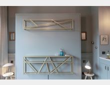 Heller Innenraum mit einer grauen Wand, an der geometrische Metallregale angebracht sind. Auf den Regalen befinden sich verschiedene Gegenstände wie ein Seifenspender und Dekorationsartikel; daneben ein kleiner Beistelltisch mit einer Tischlampe.