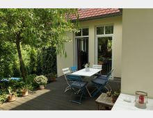 Terrasse mit Holzfußboden vor einem beigen Haus mit großen Glastüren, einem weißen Gartentisch und bunten Stühlen. Umgeben von grünen Pflanzen, Blumentöpfen und einem Baum mit herabhängenden Ästen.