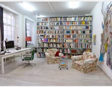 Ein heller Raum mit weißen Wänden und Holzboden, ausgestattet mit einem großen Regal voller Bücher, zwei gemusterten Sesseln und einem kleinen Beistelltisch. Links steht ein Schreibtisch mit Computer und Schreibtischlampe, daneben große Fenster mit weißen Sprossen.