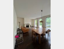 Wohnzimmer mit Holzboden, einem rechteckigen Esstisch und sechs Stühlen, einer orangefarbenen Couch sowie einem Kamin an der weißen Rückwand. Große Fenster lassen Tageslicht herein und geben den Blick auf einen Garten mit Teich frei.