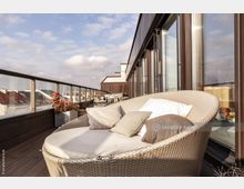 Moderne Dachterrasse mit Holzboden, Glasgeländer und Sitzmöbeln, darunter eine runde, geflochtene Liege mit Kissen. Im Hintergrund stehen kleine Tische und Pflanzenkübel, die eine Aussicht auf umliegende Gebäude bieten.