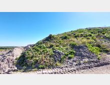 Ein Hügel mit grüner Vegetation und vereinzelten Steinen, umgeben von aufgeschütteter Erde. Im Vordergrund ein schräg aufragender Metallstab, im Hintergrund ein weiter, flacher Horizont unter einem klaren blauen Himmel.