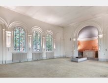 Innenraum einer ehemaligen Kapelle mit hellen Wänden und einem Boden aus cremefarbenen Fliesen. Der Raum ist geprägt von hohen Bogenfenstern mit buntem Glas und weißen Umrahmungen sowie einer Apsis mit verblassten Wandmalereien und einem steinernen Altar.