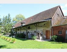 Ein ländliches Gebäude mit Fachwerk und einem Ziegeldach, umgeben von einer gepflegten Rasenfläche mit Blumentöpfen. Die Fassade ist teils aus Ziegelstein und teils verputzt, mit Holztüren und einem angrenzenden überdachten Bereich.