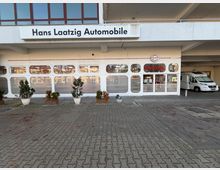 Außenansicht eines Autohauses mit der Beschriftung „Hans Laatzig Automobile“ auf einem weißen Schild. Große Glasfassade mit abgerundeten Fenstern, mehreren Topfpflanzen auf einem gepflasterten Vorplatz und ein Wohnmobil auf der rechten Seite unter einem überdachten Bereich.