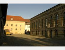 Ein gepflasterter Innenhof mit historischen Gebäuden: Links ein schlichtes Gebäude mit gelbem Putz und roten Dachziegeln, rechts ein Gebäude aus gelbem Klinker mit roten Verzierungen und hohen Rundbogenfenstern. Im Hintergrund sind weitere Gebäude sichtbar.