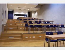 Ein Hörsaal mit ansteigenden Sitzreihen, blauen Stühlen und Holztischen, umgeben von hellgrauen Betonwänden. An der Rückwand befindet sich ein Fenster zu einem angrenzenden Raum, die Beleuchtung erfolgt durch eingelassene Deckenleuchten.