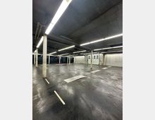 Leerer, beleuchteter Parkplatz in einem modernen, geschlossenen Parkhaus mit Betonwänden, sichtbaren Säulen und glänzendem Boden. Im Hintergrund sind Garagentore und technische Vorrichtungen zu sehen.