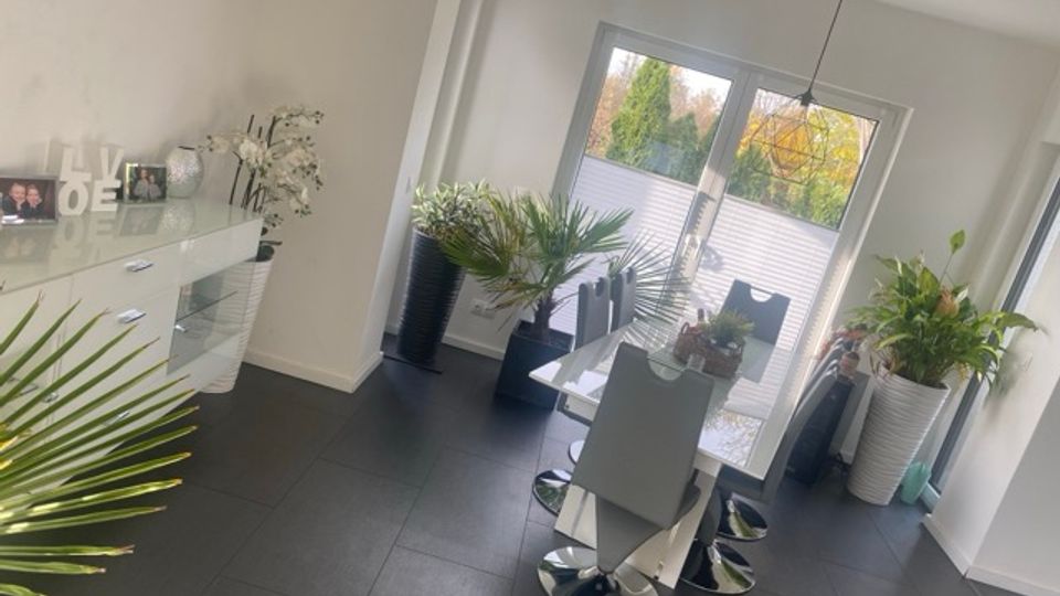 Heller, moderner Essbereich mit dunklen Fliesen, einem weißen Esstisch mit Stühlen und einer schwarzen Hängelampe. Große Fenster mit teilweiser Sichtschutzfolie öffnen den Blick ins Grün; mehrere große Topfpflanzen und ein weißes Sideboard mit Deko stehen an den Wänden.