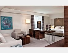 Wohnzimmerähnliches Hotelzimmer mit modernem Design, bestehend aus einer Sitzecke mit beigefarbenen Sofas, einem Couchtisch, einem Schreibtisch aus dunklem Holz mit darauf stehenden Vasen und einem Fernseher. Im Hintergrund befindet sich ein Schlafbereich mit einem Doppelbett und minimalistischem Dekor.