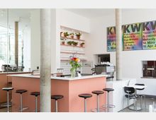 Heller, moderner Café-Innenraum mit einer Theke in Terrakottafarbe, schwarzen Hockern und bunten Blumen in einer Vase. Im Hintergrund befinden sich weiße Fliesen, Regale mit Pflanzen in Töpfen sowie Kaffeemaschinen; an der Wand hängen bunte Poster mit Veranstaltungshinweisen.