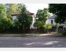 Straßenansicht mit zwei Einfamilienhäusern in einem grünen Wohngebiet. Die Häuser sind von Bäumen und Sträuchern umgeben, und ein gepflasterter Gehweg verläuft entlang der Straße mit Kopfsteinpflaster.