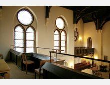 Innenansicht einer historischen Kirche mit hohen Buntglasfenstern in Spitzbogenform. Sicht auf eine hölzerne Empore, Sitzbänke und einen kleinen Bereich mit einfachen Stühlen und Tischen.