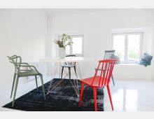 Heller Raum mit minimalistischer Einrichtung, bestehend aus einem runden weißen Tisch mit filigranen Metallbeinen, umgeben von farbigen Stühlen (grün, rot und blau). Im Hintergrund befinden sich zwei Fenster mit Sitzbank, dekoriert mit Kissen.