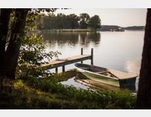 Ein ruhiger See mit einem hölzernen Steg, an dem ein kleines grünes Ruderboot befestigt ist. Im Hintergrund sind Bäume und Schilfgruppen am Ufer zu sehen.