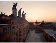 Backsteinindustriegebäude mit markanten Schornsteinen auf dem Dach, aufgenommen bei Sonnenuntergang. Im Hintergrund sind weitere Gebäude sowie ein flacher, weitläufiger Hof und der leicht bewölkte Himmel zu sehen.