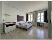 Helles Schlafzimmer mit Holzfußboden, einem Doppelbett, großen weißen Sprossenfenstern mit blickdichten Vorhängen und einer langen weißen Kommode. Links steht ein Wickeltisch, an der Wand hängt ein abstraktes Gemälde.