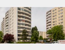 Mehrstöckige Wohngebäude mit Balkonen in einer städtischen Umgebung, umgeben von Bäumen und parkenden Autos. Im Vordergrund befinden sich grüne Sträucher und ein Baum, die den Eingang zu einem der Gebäude säumen.