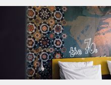 Bunt gestaltete Wand mit großflächigem Blumenmuster im Retro-Stil und der Aufschrift „the 70s“. Im Vordergrund sind ein gelbes Kopfteil eines Bettes und weiße Kissen zu sehen, daneben eine chromfarbene Hängelampe.