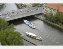 Blick auf einen Fluss mit zwei Passagierschiffen, die daran vorbeifahren, und einer Brücke, die darüberführt. Am Ufer befinden sich Bäume, ein Gehweg und Gebäude mit roten Dächern.