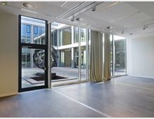 Moderner Innenraum mit großen Glasfenstern, die Blick auf einen Innenhof mit geometrischer Metallskulptur ermöglichen. Die Decke ist mit integrierten Lampen und Lüftungselementen gestaltet, Vorhänge hängen teilweise vor den Fenstern.