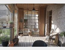 Überdachte Veranda mit Holzboden und einer Wand aus unverputzten Ziegeln; im Hintergrund eine große Fensterfront mit Metallrahmen. Der Bereich ist mit Korbmöbeln, Pflanzen und rustikalen Dekorationen ausgestattet.