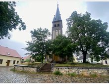 Kirchplatz mit einer steinernen Kirche im Zentrum, die einen hohen Turm mit Spitzdach und Uhr besitzt. Umgeben von Kopfsteinpflaster, Bäumen und eingeschossigen Gebäuden mit Satteldächern.