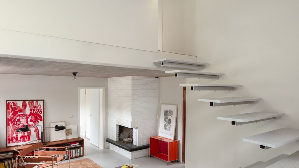Minimalist split-level living room with white walls, a floating staircase leading to an upper opening, and a white brick corner fireplace. The lower seating area includes modern chairs, a tan rug, low shelving with framed artwork, and a small red cabinet.