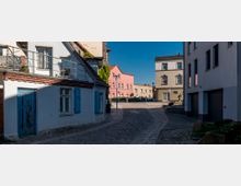Kopfsteinpflasterstraße in einem Wohngebiet mit mehrfarbigen Gebäuden; links ein blau-weißes Haus mit blauen Fensterläden und darüber eine Terrasse, rechts moderne Gebäude mit Garagen. Im Hintergrund stehen beige und rosa Wohnhäuser.