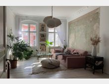 Ein helles Wohnzimmer mit Holzdielenboden, einem großen Sprossenfenster und einer historischen Stuckdecke. Die Einrichtung umfasst ein rosa Sofa, einen runden Pouf, einen Glastisch und zahlreiche Zimmerpflanzen sowie eine halb verputzte Wand und botanische Wandbilder.