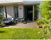 Terrasse eines modernen Wohnhauses mit Holzboden, darauf eine Sitzecke mit Tisch, Stühlen und einer Sonnenliege. Umgeben von Pflanzen in Töpfen und angrenzend an einen sonnigen Rasen.