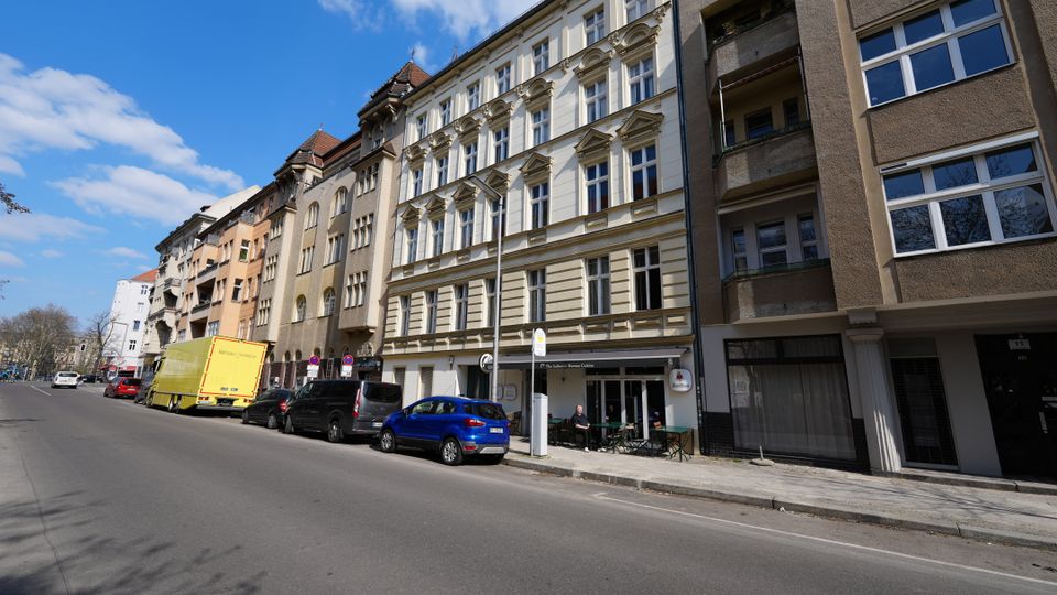 Straßenansicht einer städtischen Wohnhauszeile mit mehrstöckigen Altbaufassaden, darunter ein helles Gebäude mit reich verzierten Fensterrahmen, neben moderneren Häusern mit Balkonen. Entlang der breiten Fahrbahn stehen geparkte Autos und ein Lkw; am Erdgeschoss sind Schaufenster und ein kleiner Außenbereich erkennbar, darüber blauer Himmel mit Wolken.