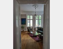 Wohnzimmer mit Holzboden, einer gläsernen Couchtisch auf einem gemusterten roten Teppich und Sitzmöbeln in schwarz-weißem Rautenmuster. Die weißen Wände sind mit hohen Fenstern, einer Deckenrosette und einem Kristallkronleuchter verziert; an der Wand hängt ein Gemälde, und eine Zimmerpflanze steht in der Ecke.