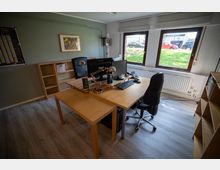 Ein Büro mit einem großen Schreibtisch aus hellem Holz, ausgestattet mit Computermonitoren, Tastaturen, Tischlampen und Bürostühlen. Drei große Fenster lassen Tageslicht herein und bieten Blick auf eine Grünfläche und parkende Autos; Regale und Wanddekoration ergänzen den Raum.
