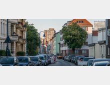 Wohnstraße mit parkenden Autos auf beiden Seiten, flankiert von Altbauten mit verzierten Fassaden links und moderneren Gebäuden rechts. Im Hintergrund sind Bäume, Baustellenabsperrungen und ein Hochhaus zu sehen.