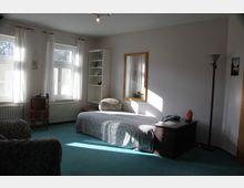 Ein Schlafzimmer mit Teppichboden in Türkis, einem Einzelbett, einem kleinen Nachttisch und einer Kommode. Das Zimmer hat zwei große Fenster mit weißen Vorhängen, ein Bücherregal, einen großen Spiegel an der Wand und eine Stehlampe in der Ecke.