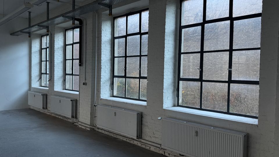 Innenraum eines industriellen Loft- oder Bürogebäudes mit weiß gestrichenen Ziegelwänden und großen, schwarz gerahmten Sprossenfenstern entlang der Wand. Unter den Fenstern sind mehrere Heizkörper angebracht; an der Decke verlaufen sichtbare Kabeltrassen und Leitungen.