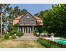 Zweistöckiges Wohnhaus mit dunkler Fassade und rotem Ziegeldach, umgeben von Bäumen. Im Vordergrund befinden sich ein gepflegter Garten, eine Terrasse mit Sitzmöbeln und ein Pool mit Holzumrandung.