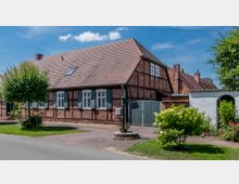 Traditionelles Fachwerkhaus mit rotem Ziegeldach und blauen Fensterläden, umgeben von einem gepflegten Garten mit Blumen und Bäumen. Im Vordergrund ist eine gepflasterte Einfahrt sowie ein kleiner grüner Vorgarten zu sehen.