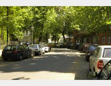Straßenansicht mit geparkten Autos auf beiden Seiten, umgeben von hohen, grünen Bäumen, die Schatten auf die Fahrbahn werfen; im Hintergrund sind Wohngebäude und ein rotes Backsteinhaus mit großen Fenstern zu sehen.