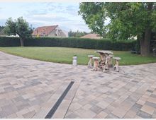 Ein gepflasterter Hof mit hellen und dunklen Steinen grenzt an eine gepflegte Rasenfläche, die von einer Hecke und einem großen Baum gesäumt wird. Im Vordergrund steht eine rustikale Sitzgarnitur aus verwittertem Holz, und im Hintergrund sind Häuser sowie ein Gebäude im Bau zu sehen.