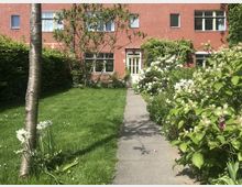 Roter Wohnhausfassade mit weißen Fenstern, umgeben von einem gepflegten Garten mit grünem Rasen, Blumenbeeten und Sträuchern. Ein schmaler gepflasterter Weg führt durch den Garten zu einer gelben Eingangstür.