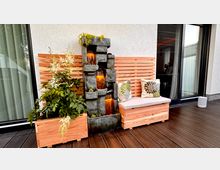 Dekorative Sitzecke auf einer Terrasse mit Holzboden, bestehend aus einer Holzbank mit Kissen, flankiert von einem bepflanzten Blumenkasten und einem kleinen Wasserfall aus Stein. Im Hintergrund eine Holzwand und eine verglaste Schiebetür, die ins Gebäude führt.
