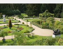 Ein symmetrischer, gepflegter Garten mit geschwungenen Kieswegen, geschnittenen Hecken, Blumenbeeten und einer überdachten Laube mit Rankpflanzen. Im Vordergrund sind Pflanzen in Kübeln, eine steinerne Vase und eine Bronzestatue auf einem Sockel zu sehen, umgeben von dichter Vegetation.