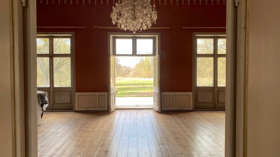 Großer historischer Saal mit Holzdielenboden, dunkelroten Wänden und Stuckfries an der Decke, in der Mitte ein Kristallkronleuchter. Drei hohe Fenstertüren, die mittlere geöffnet, führen auf eine Grünfläche im Außenbereich.